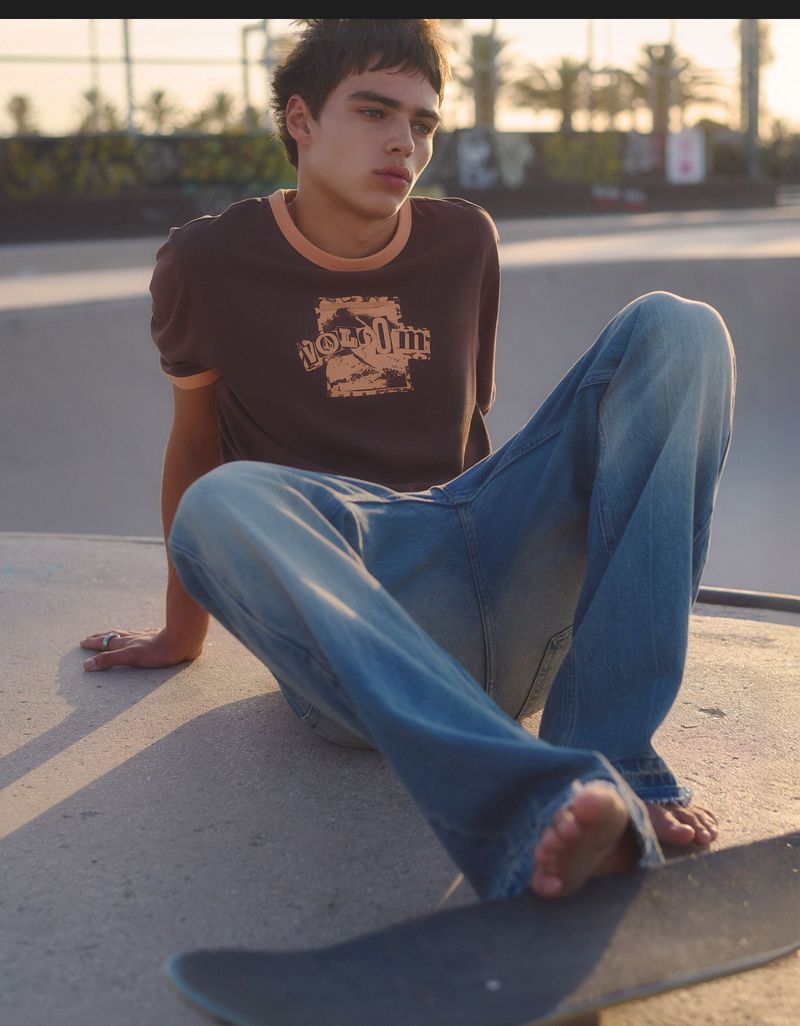 Jon Payeras sitting barefoot at a skatepark wearing a brown graphic ringer tee and baggy jeans next to a skateboard.
