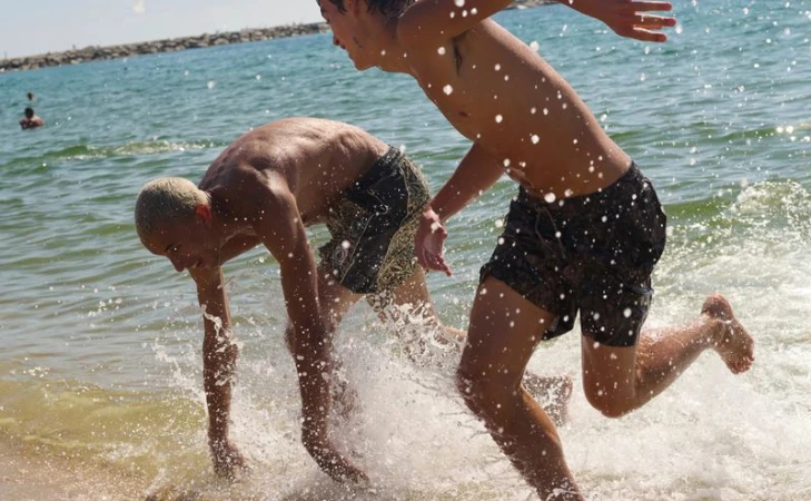 Two young men running and splashing through shallow water on a sunny beach in swim trunks.