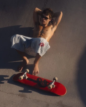 Model Jon Payeras relaxes shirtless in baggy denim shorts on concrete next to a red skateboard in direct sunlight.