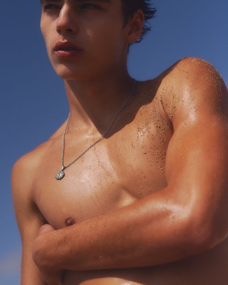 Shirtless young man with sand on his sunlit shoulder wearing a silver pendant necklace against a clear blue sky.
