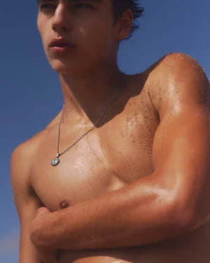 Shirtless young man with sand on his sunlit shoulder wearing a silver pendant necklace against a clear blue sky.