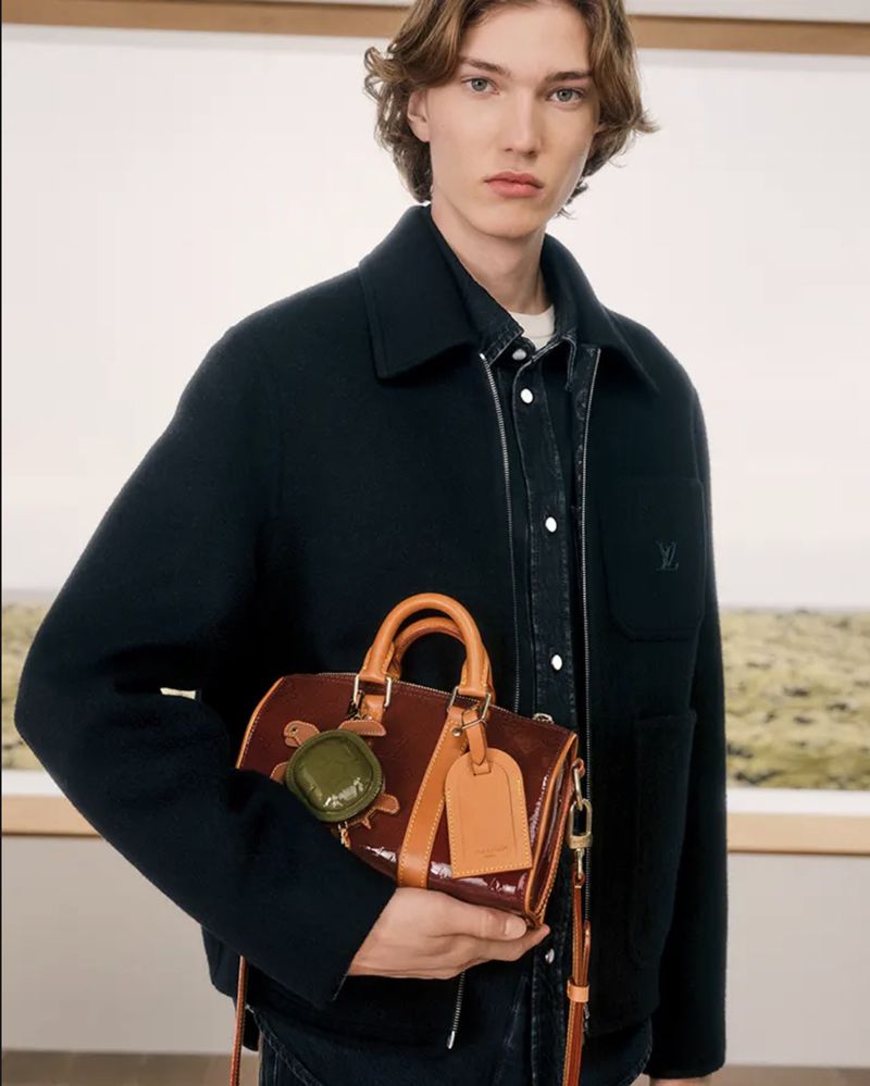Model wearing a dark wool zip jacket holding a glossy burgundy and tan mini handbag with bag charms.