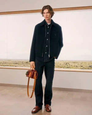 Model in dark zip jacket and trousers holding a brown leather cylinder bag in front of a wide horizontal window.