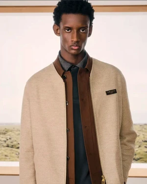 Black male model wearing a beige zip jacket layered over a brown collared shirt standing before a framed landscape backdrop.