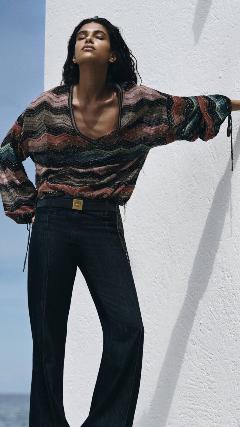 Model Tamar David posing in a multicolor chevron knit top and dark wide-leg denim against a white textured wall.