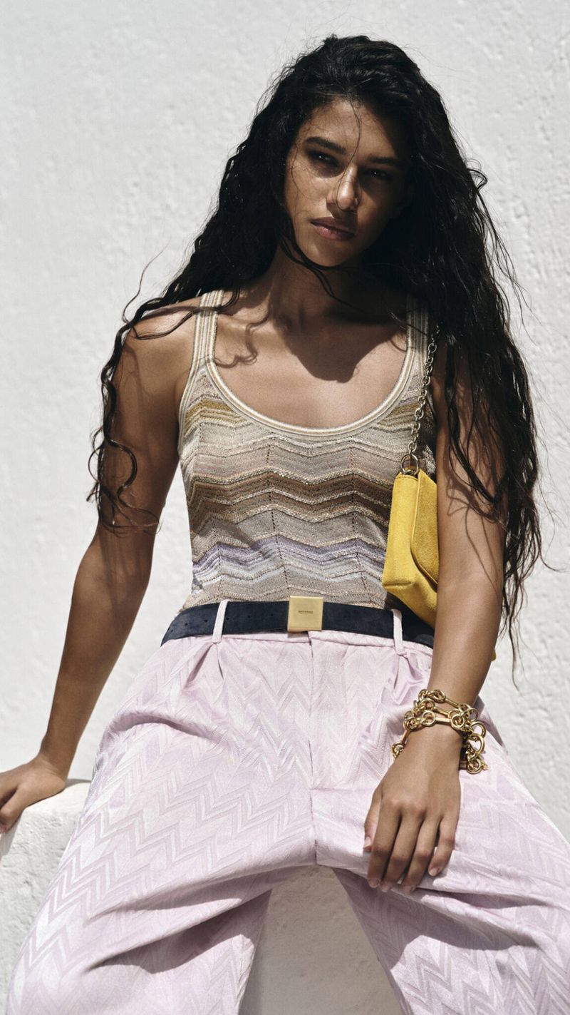 Model Tamar David wearing a chevron knit top, wide-leg patterned trousers, and a yellow bag against a textured white wall.