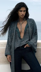 Model Tamar David wearing a textured blue knit cardigan and dark denim jeans outdoors with the sea in the background.