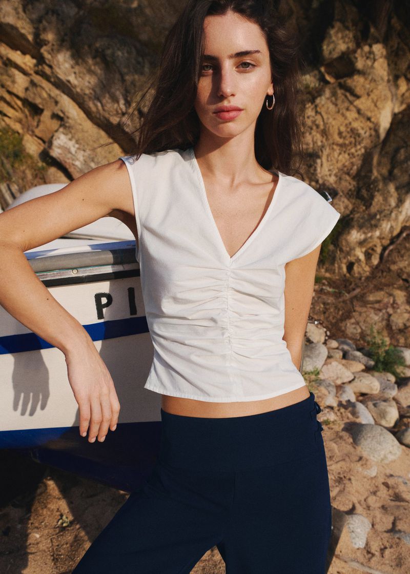 Model wearing a white ruched sleeveless V-neck top and dark trousers leaning against a white boat beside a rocky hill.