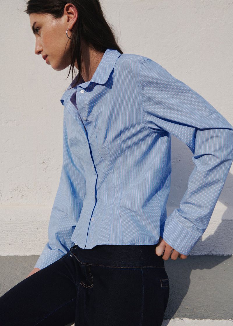 Woman in a light blue striped button-up shirt and dark jeans posing against a white textured wall in direct sunlight.