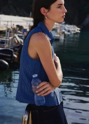 A woman in a blue pinstriped sleeveless top holding a water bottle with a harbor and boats visible in the background.