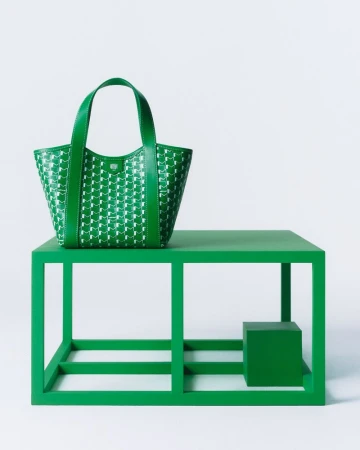 Green leather tote bag with geometric design sits atop a matching green, open-frame table against a plain white backdrop.