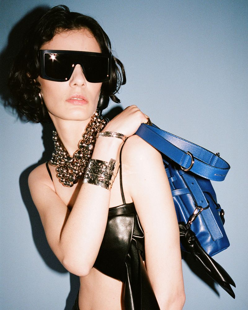 Model wearing oversized black shield sunglasses, metallic ball jewellery, and a black leather top with a royal blue shoulder bag.