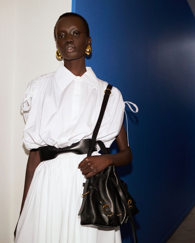 Model wearing a white shirt and skirt holding a black handbag against a wall
