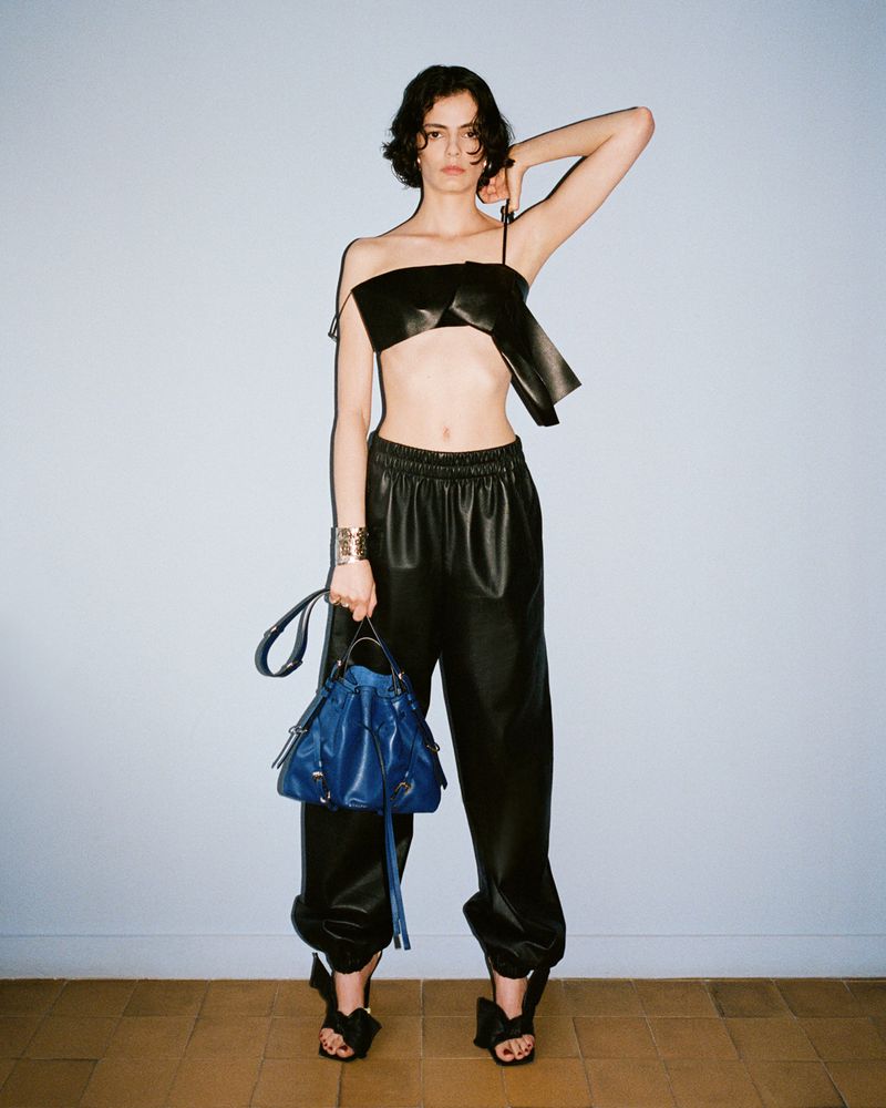 Model wearing a black leather bandeau and trousers holding a blue handbag against a plain grey studio wall.