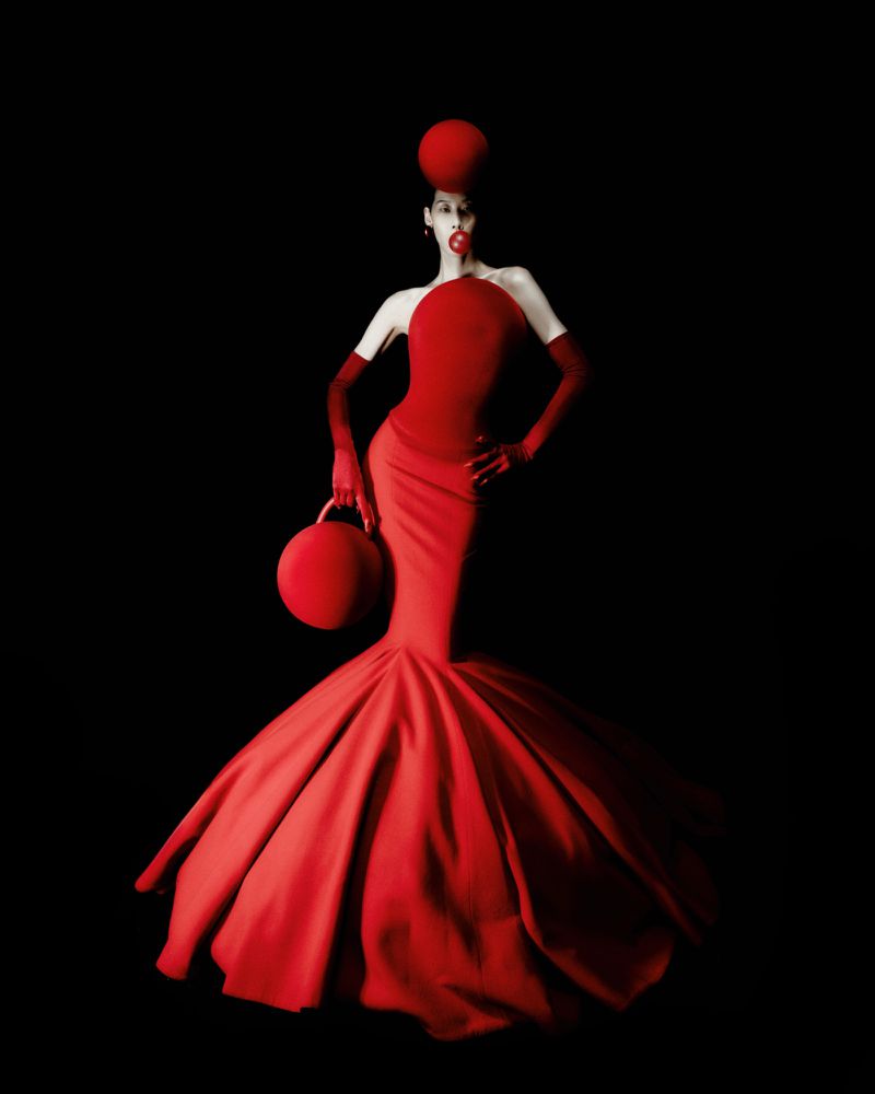 Model posing in a sculptural red mermaid gown with matching gloves, a spherical red headpiece, and circular handbag against black.