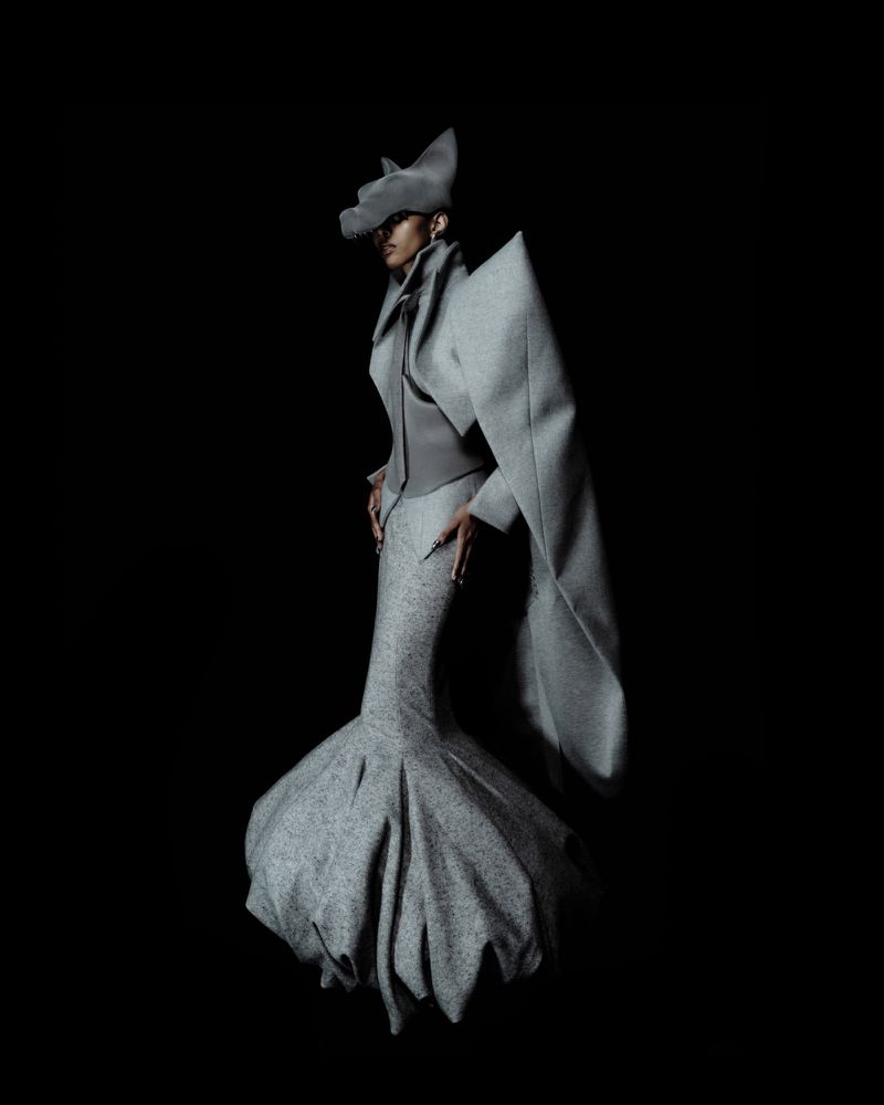Model wearing a sculptural grey Robert Wun couture outfit with an exaggerated silhouette against a solid black background.
