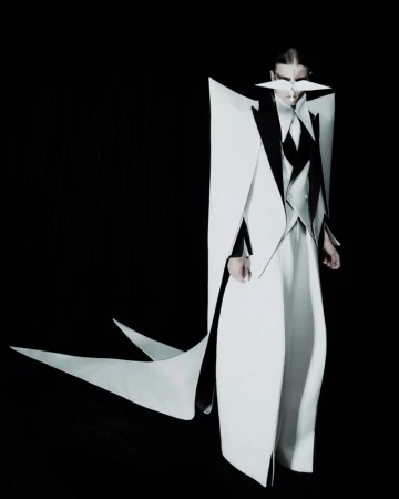 Model wearing an avant-garde sculptural white and black coat with sharp geometric shoulders and face accessory against a black background.