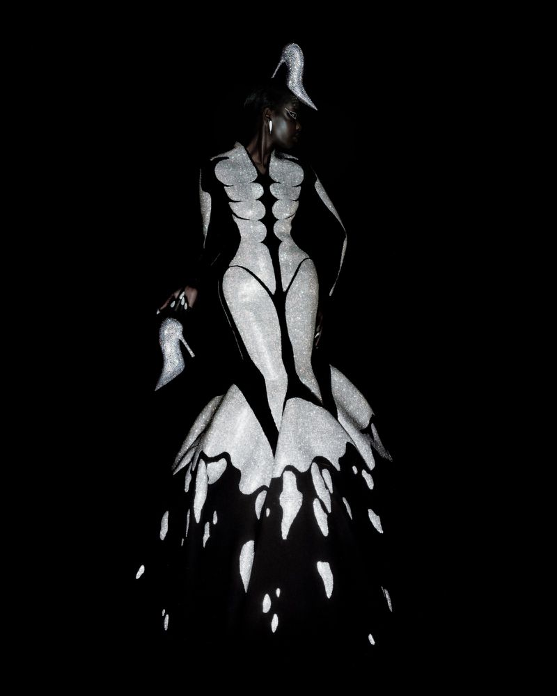 Model wearing a surreal black and sparkling silver Robert Wun couture gown with high heel shoe accents against a black background.