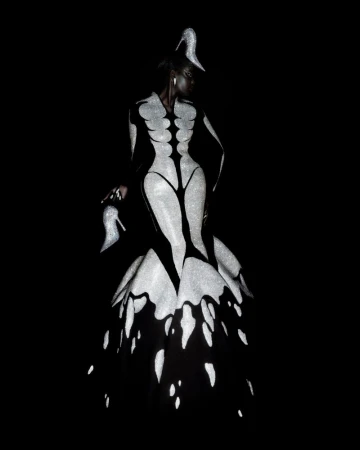 Model wearing a surreal black and sparkling silver Robert Wun couture gown with high heel shoe accents against a black background.