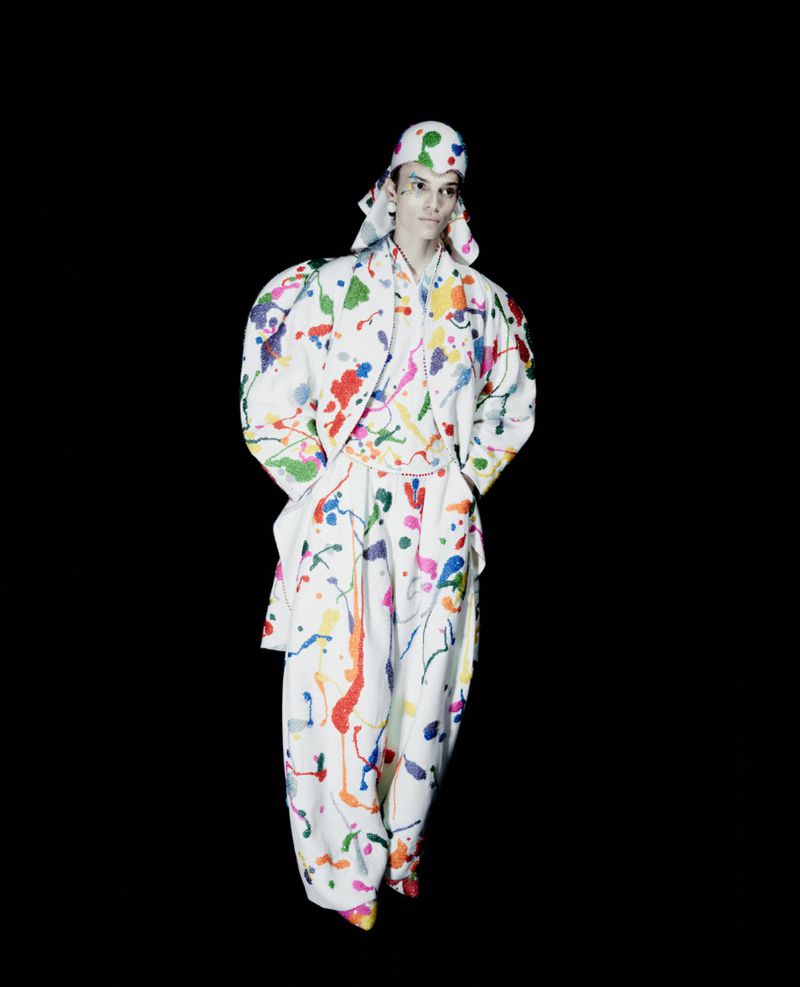 Fashion model wearing a white paint-splattered voluminous suit with matching draped headwear against a pitch black background.