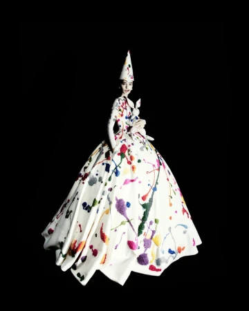 Model wearing an off-white paint-splattered couture ballgown and matching cone hat while holding a stuffed rabbit against a black backdrop.
