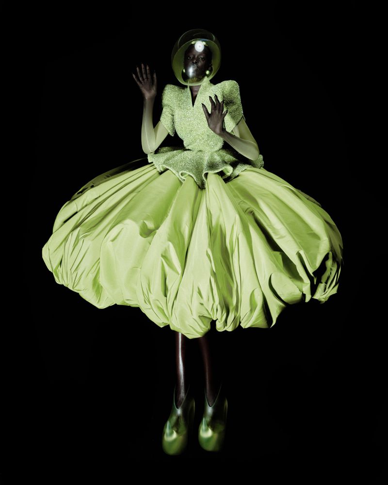 A Black model wearing a sculptural lime green puff dress and clear dome helmet floating against a black seamless backdrop.