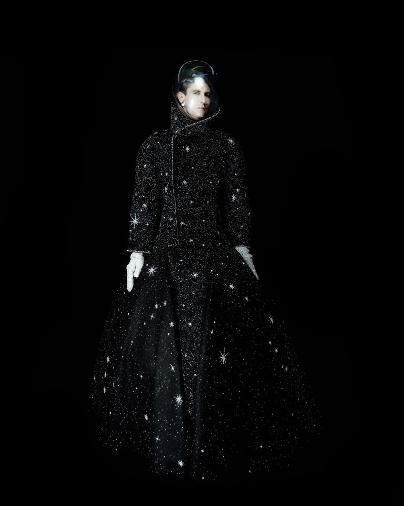 Model wearing an embellished black starry haute couture gown with a clear helmet hood and white gloves against a black background.