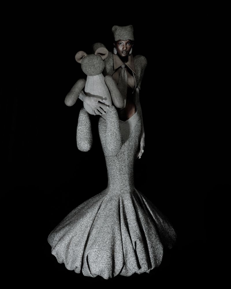 Model wearing a sculptural grey wool mermaid gown and holding a matching fabric teddy bear against a dark black background.