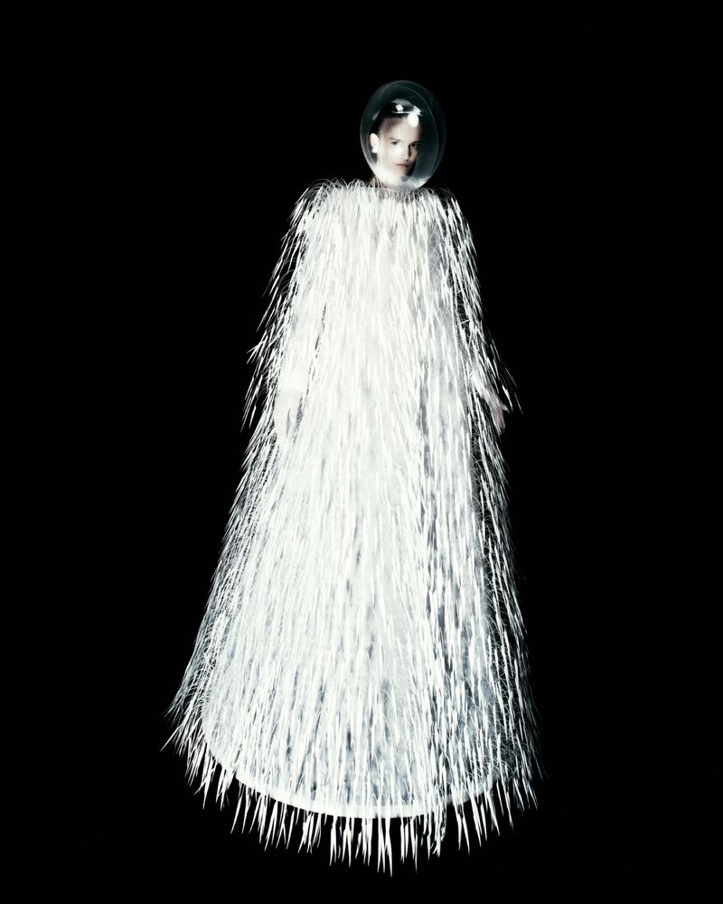A model in a floor-length textured white fringe gown wearing a clear spherical helmet against a solid black background.