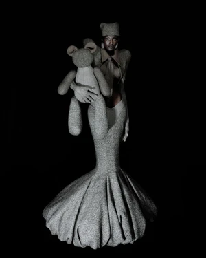 Model wearing a sculptural grey wool mermaid gown and holding a matching fabric teddy bear against a dark black background.