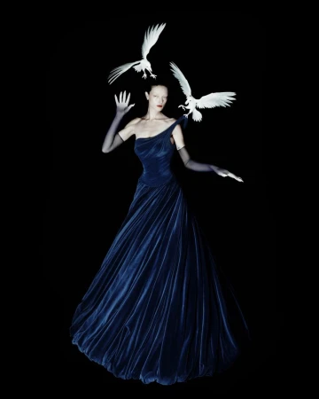 Model in a midnight blue velvet gown and sheer black gloves surrounded by sculpted white birds against a solid black background.