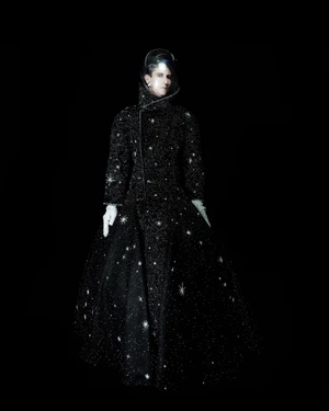 Model wearing an embellished black starry haute couture gown with a clear helmet hood and white gloves against a black background.