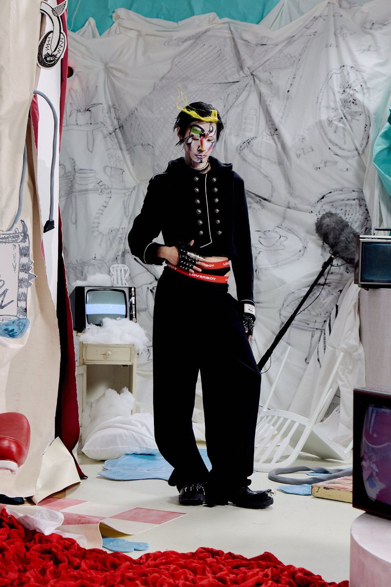 Model with avant-garde facial makeup wearing a black cropped military jacket and wide trousers on an illustrated studio set.