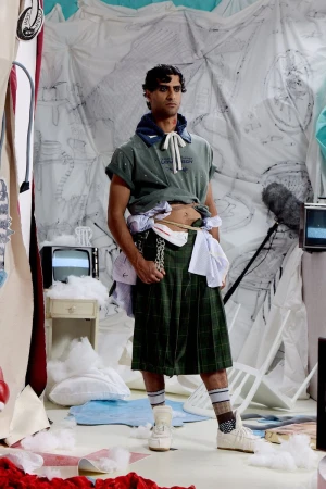 Model standing in a green plaid kilt and rolled-sleeve t-shirt on a set-dressed studio stage for Charles Jeffrey Loverboy.