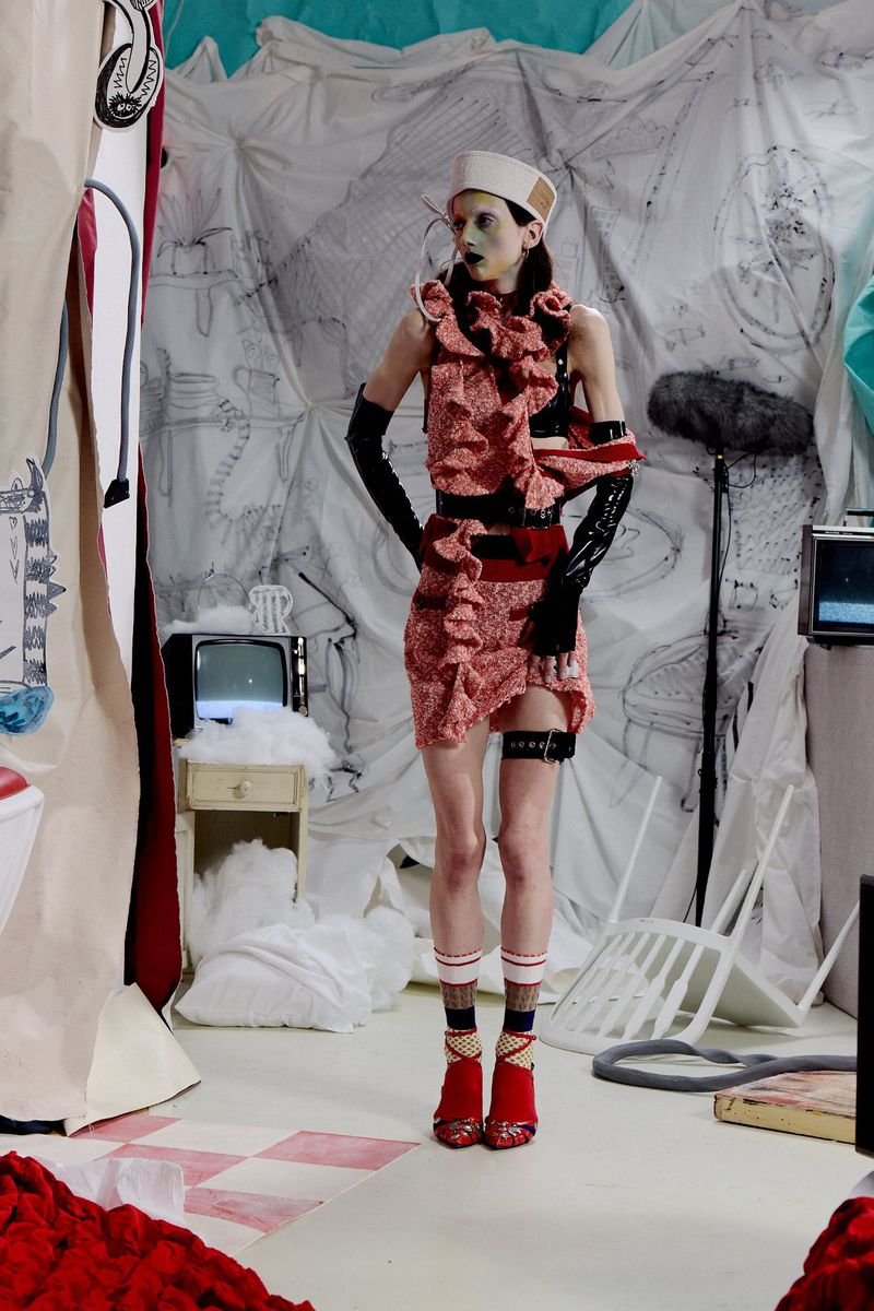 Model wearing a red ruffled dress, sailor cap, and black gloves in an eclectic set-dressed studio environment.