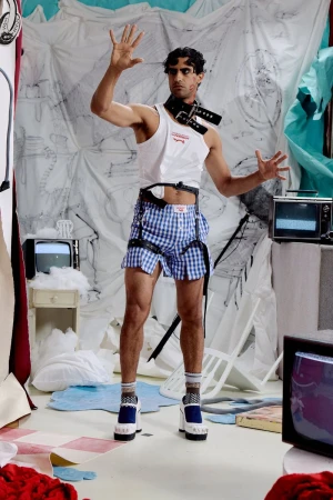 Model wearing a white tank top, blue gingham shorts, and black leather harness posing on an illustrated studio set.