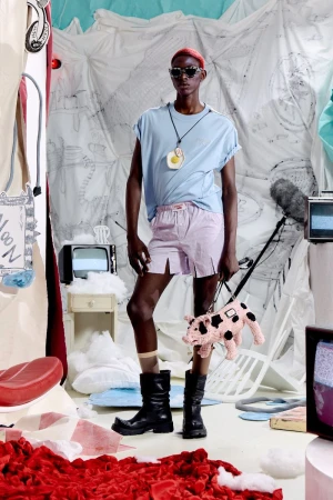 Model Quadri Ajao wears a light blue tee, lilac shorts, sunglasses, and black boots while holding a pig handbag for Charles Jeffrey Loverboy.