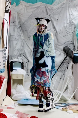 Model wearing an avant-garde layered graphic knit poncho, animal-ear headwear, and platform heels on a set-dressed studio stage.