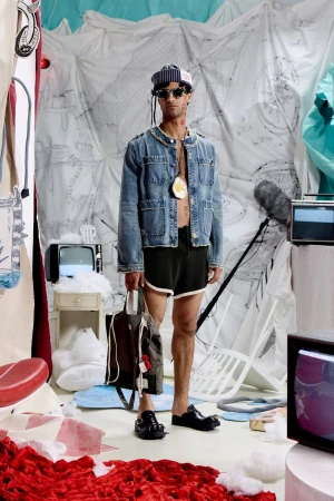 Model Quadri Ajao in a denim jacket, black shorts, and sailor cap holding a tote bag on an eclectic set with vintage televisions.