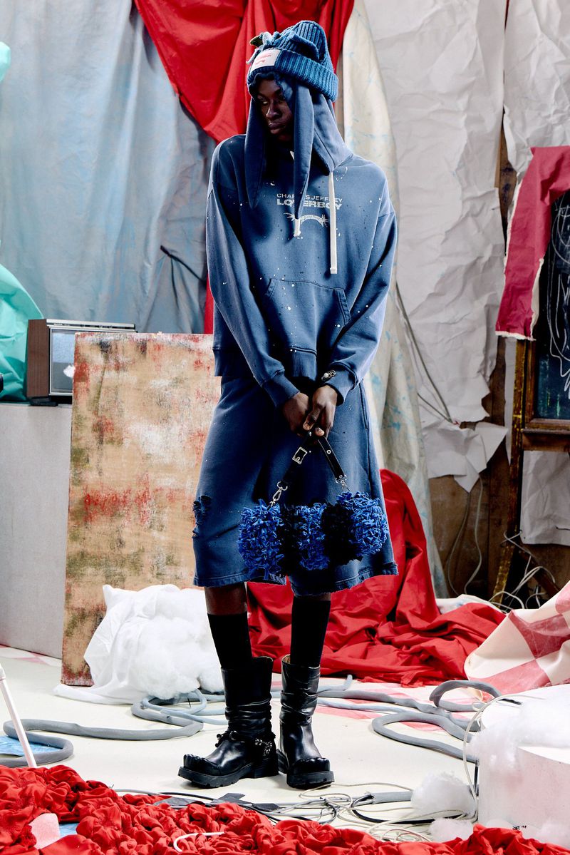 Model Quadri Ajao standing in a blue graphic hoodie, matching shorts, knit ear beanie, and black chunky boots on a styled studio set.