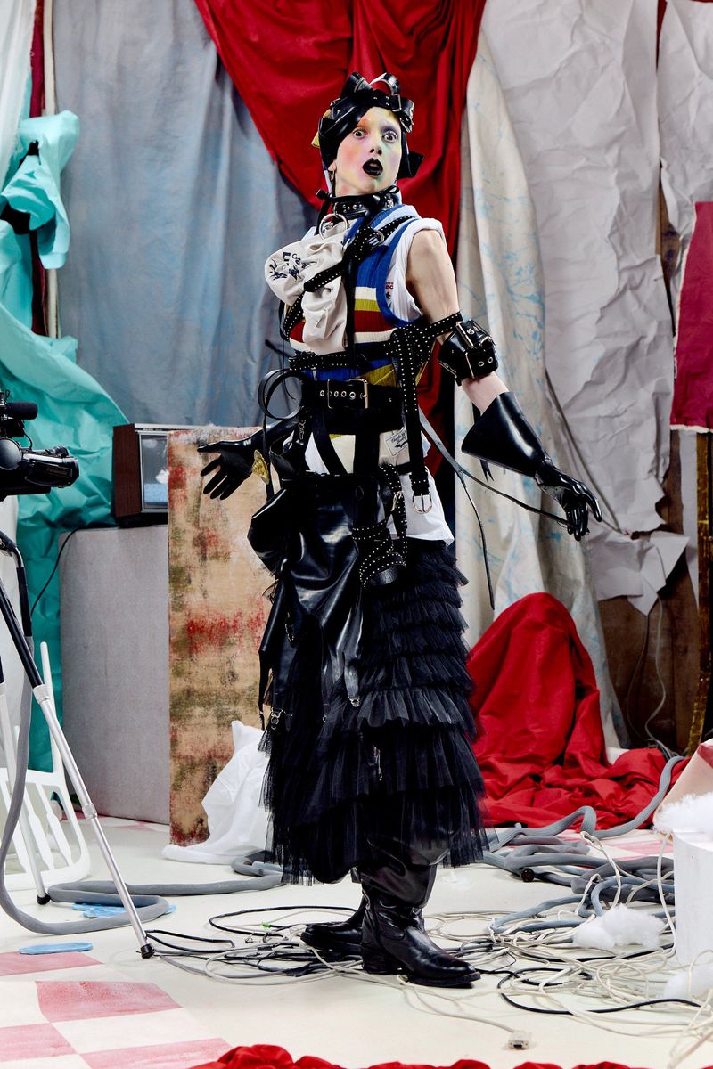 Fashion model in an avant-garde layered black tulle skirt and harness standing in a draped fabric studio set.