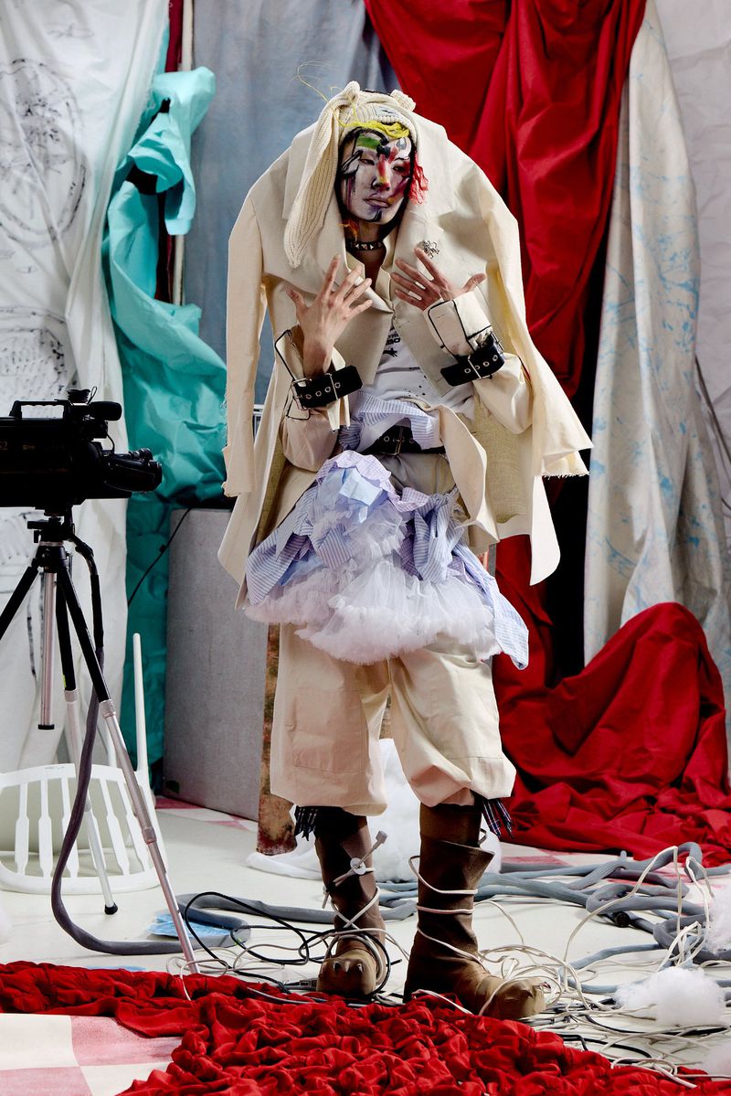 Model in an avant-garde cream jacket, painted makeup, and ruffled skirt posing on a studio set with red drapery and a camera on a tripod.