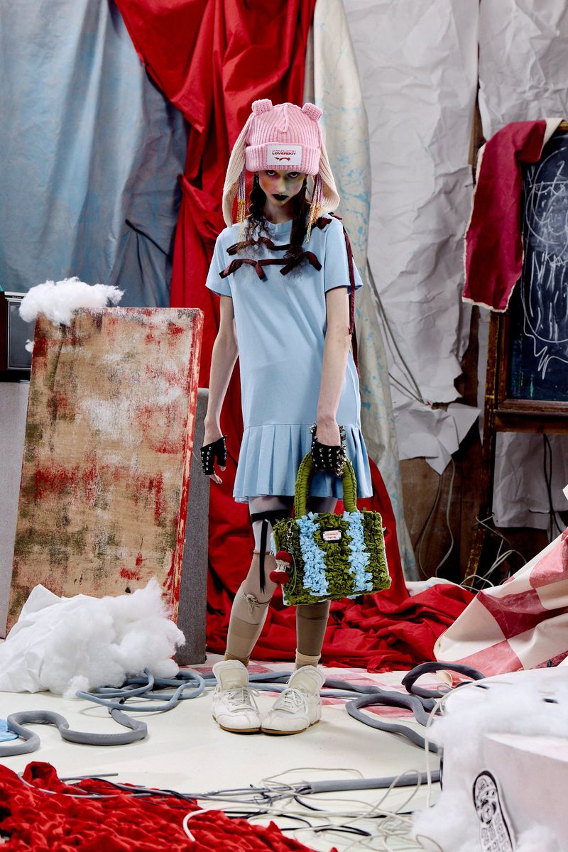 Model in a light blue pleated dress and pink knit animal-ear beanie holding a textured handbag on a chaotic studio set.