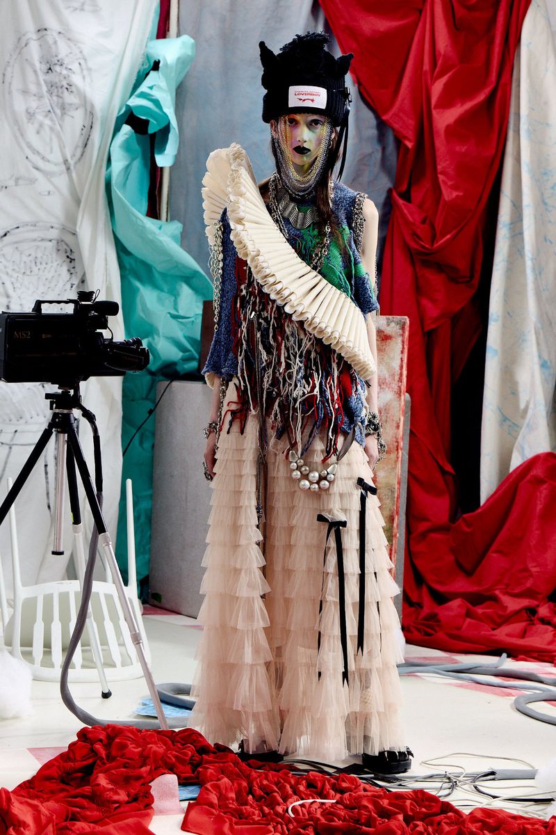 Model Zara Sands wearing an avant-garde tiered tulle skirt and cat-ear beanie on a studio set with draped red fabric.