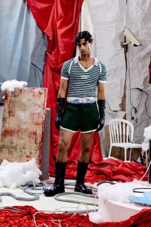 Fashion model posing in a green striped knit top, dark green shorts, black leather gloves, and tall boots on a red draped studio set.