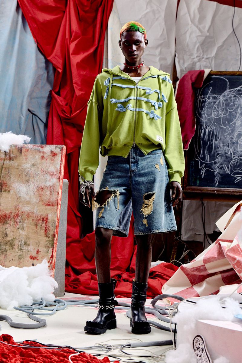 Model Quadri Ajao wearing an embellished green hoodie, distressed denim shorts, and black boots in a studio set.