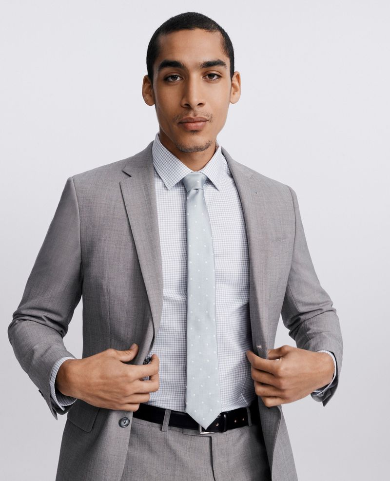 Man in a tailored light grey suit, patterned dress shirt, and light tie posing against a white studio background.