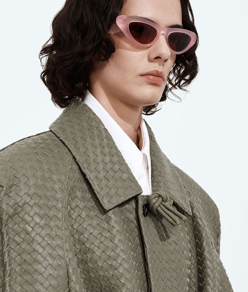 Model wearing translucent pink cat-eye sunglasses and an olive woven leather jacket against a seamless white studio background.
