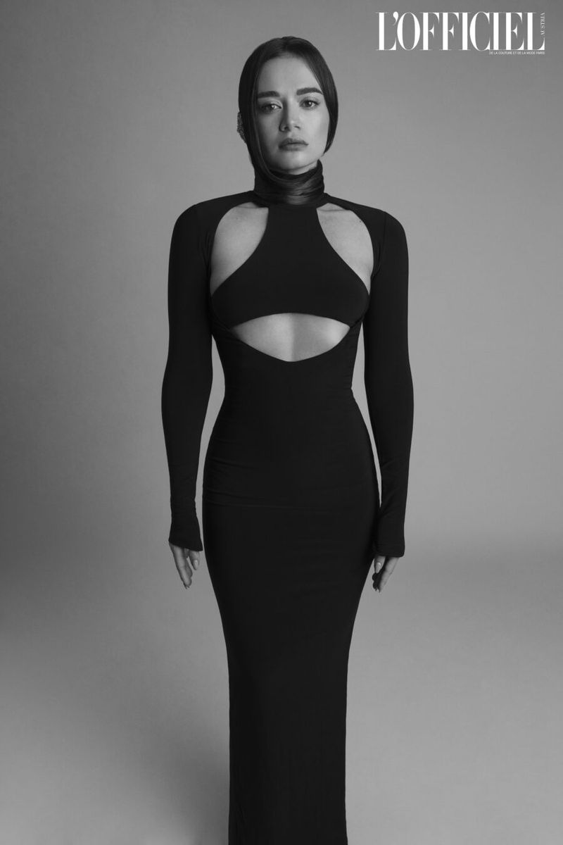 Elegant woman in a black, cutout dress posing in a studio, showcasing fashion and style in a monochromatic setting.