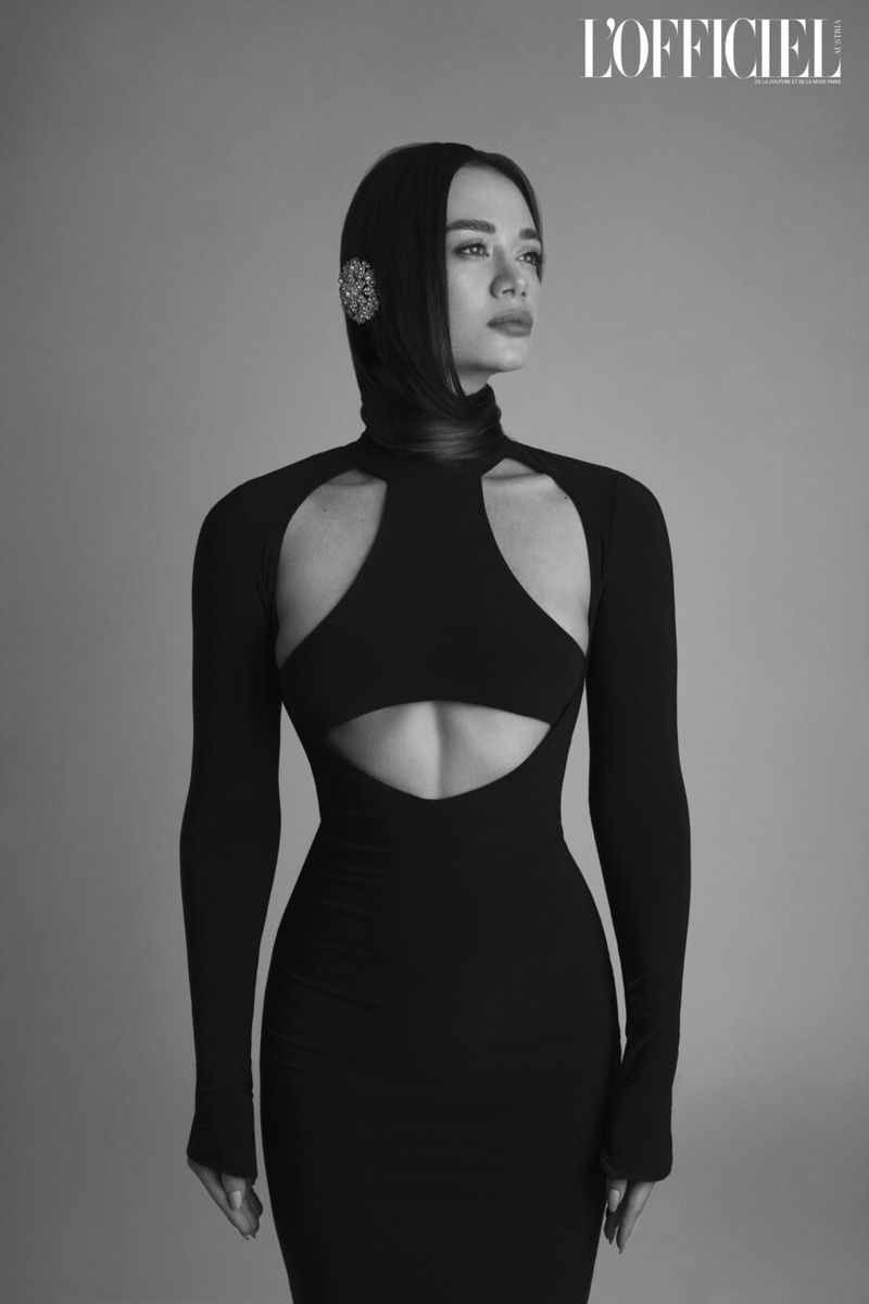 Monochrome portrait of a woman in a cutout black dress and head covering, gazing off-camera against a neutral background, with L'Officiel logo.